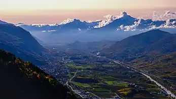 Mountainous landscape of a wide valley seen from above during a sunset. You can see several villages, fields and a river. In the distance, the mountains are in the clouds.