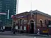 A red bricked cube shaped building with a rectangular, dark blue sign reading "PLAISTOW STATION" in white letters and people walking on the pavement in front