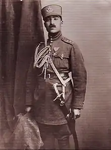 Sepia photograph of Mahmud Khan Pulādeen standing in a military uniform, there's a cloth on the left-hand side of the wall behind him