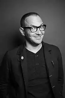 Black-and-white photograph of a man with short hair and black glasses, wearing a face microphone.