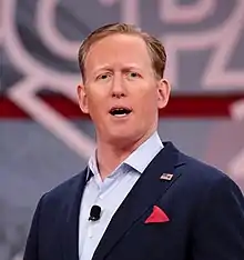 A bust photograph of a white man; he is wearing a blue collared shirt and jacket while looking at the camera and speaking