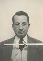 Mug shot of a man with glasses in suit and tie. The number O-15 appears in front of him.