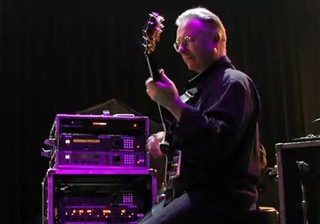 An older man from the side, holding a guitar with an amp to his left under a purple spotlight