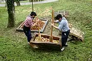 Two individuals using large wooden mallets to pound apples in a traditional wooden trough in the Basque Country.