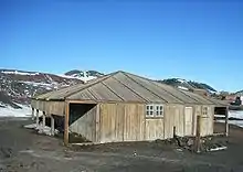 Wooden structure with door and two small windows. To the left is an open lean-to. In the background are partly snow-covered mountains