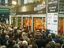 Small building with glass walls, with signs on top and spectators outside