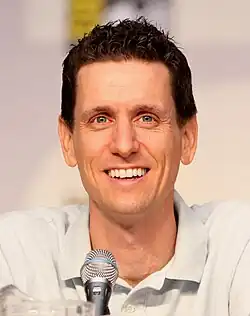 A man with brown hair, leans forward slightly while speaking into a microphone.