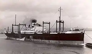 Starboard side of a large black ship; she has a single funnel and is being towed by a tug.