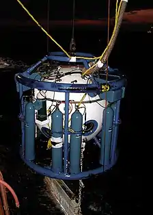 Night view of a white spherical pressure chamber in a blue pipe frame supporting several blue bulk gas storage clinders, suspended over the water by cables. The bell umbilical is visible at the top and a ballast weight can be seen below at the water surface